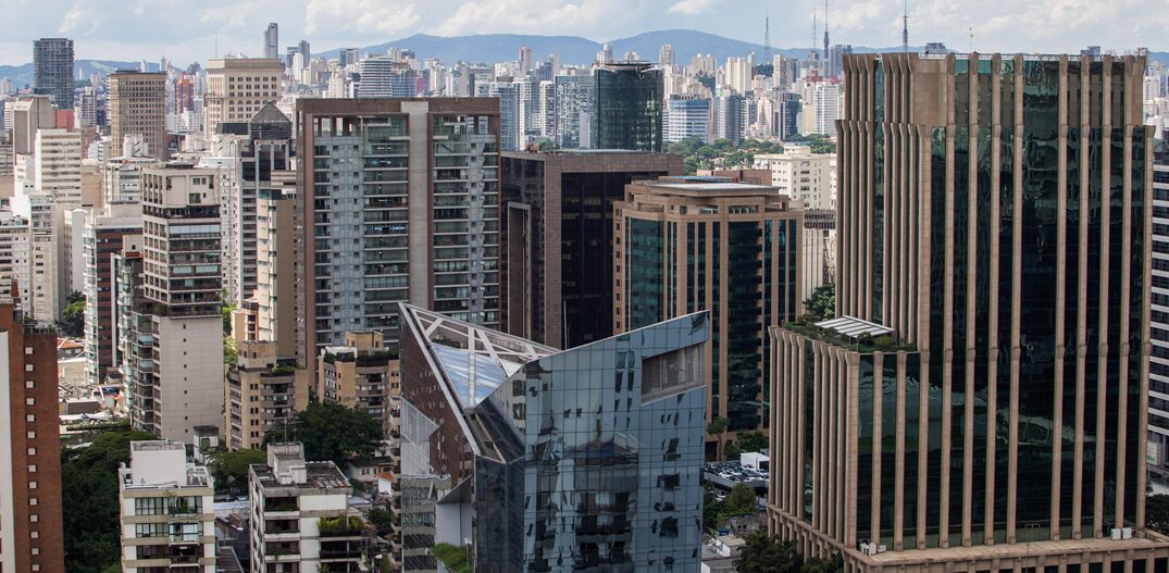 Cidade de São Paulo vista de longe, vários prédios modernos aparecem em primeiro plano e ao fundo podemos ver montanhas ao longe, tudo sob um céu azulado, porém com muitas núvens. Foto: FOTO TIAGO QUEIROZ / ESTADÃO  