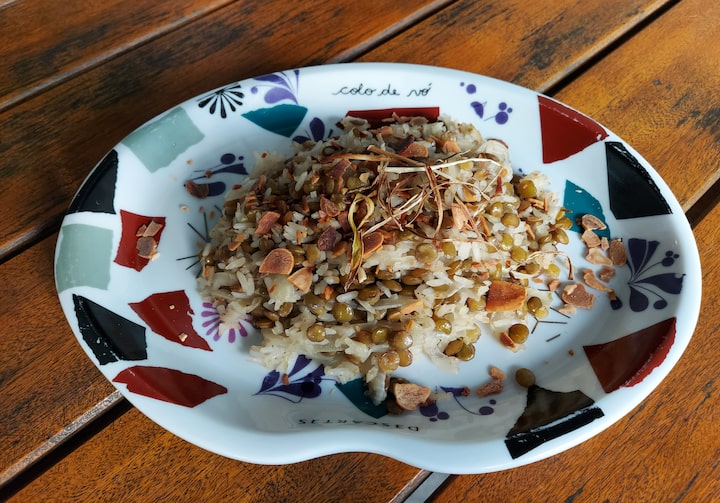 Sobre uma mesa de madeira, está um prato colorido de louça e com bordas ligeiramente altas. Dentro, está uma porção de arroz com lentilhas, cebola e amêndoas.