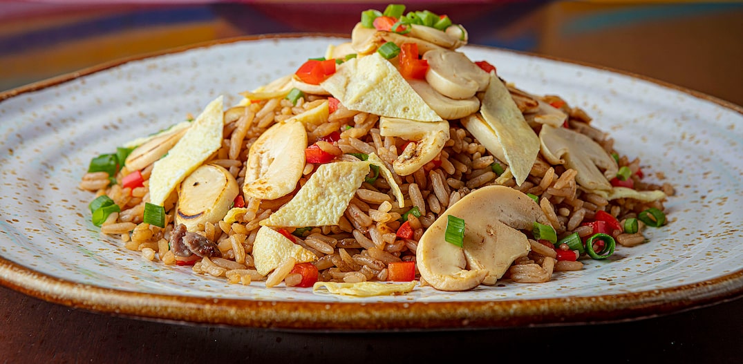 Sobre um prato raso de louça está uma porção de arroz com cogumelos fatiados por cima, cenouras picadas e temperos frescos verdes. Foto: Luis Vinhão