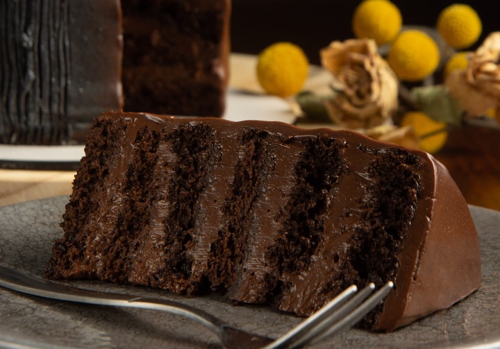 Um pedaço de bolo de chocolate está sobre um prato cinza, com um garfo ao lado. Ao fundo, temos o bolo do qual foi retirada a fatia e decoração de bolinhas amarelas e flores ao lado.