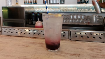 Sobre uma bancada de bar clara, está um copo transparente contendo um canudo branco com azul e o drinque Mykonian, uma versão de soda italiana com xarope de cereja. Foto: Giulia Howard/Estadão
