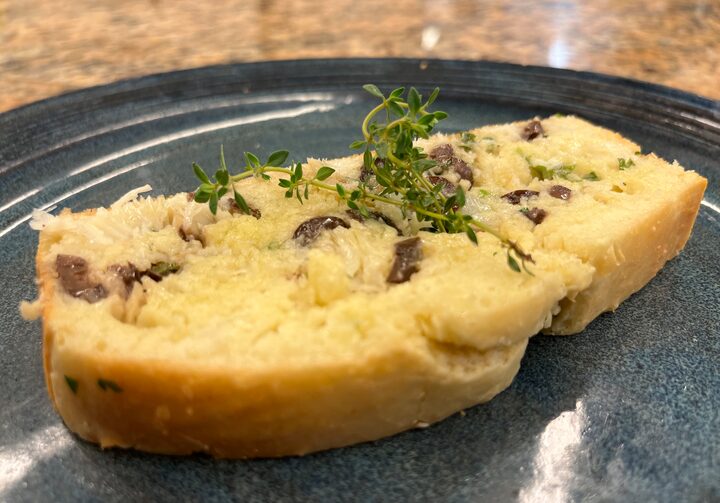 Em um prato de louça azul-escura, uma fatia da torta de bacalhau, que apresenta azeitonas e bacalhau no recheio, servida com um ramo de tomilho fresco por cima.