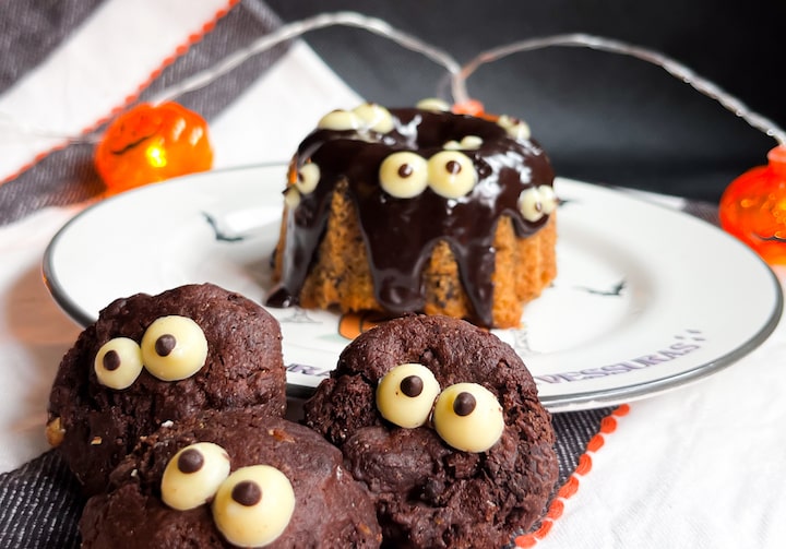 Cookies de chocolate com olhinhos feitos de chocolate branco e chocolate meio amargo. Ao fundo, há um prato com bolo de cenoura com calda e os olhinhos de chocolates