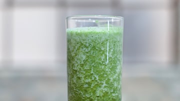 Suco verde de couve, abacaxi, chia, gengibre e hortelã dentro de copo de vidro transparente. O objeto está sobre superfície plana de madeira clara. Foto: Vitória Magalhães | Anhembi Morumbi