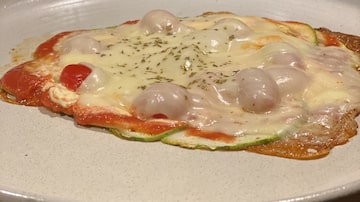 Sobre um prato de louça de cor cinza clara, uma versão de pizza de abobrinha preparada sobre crosta de queijo crocante. Foto: Gabriela Junqueira