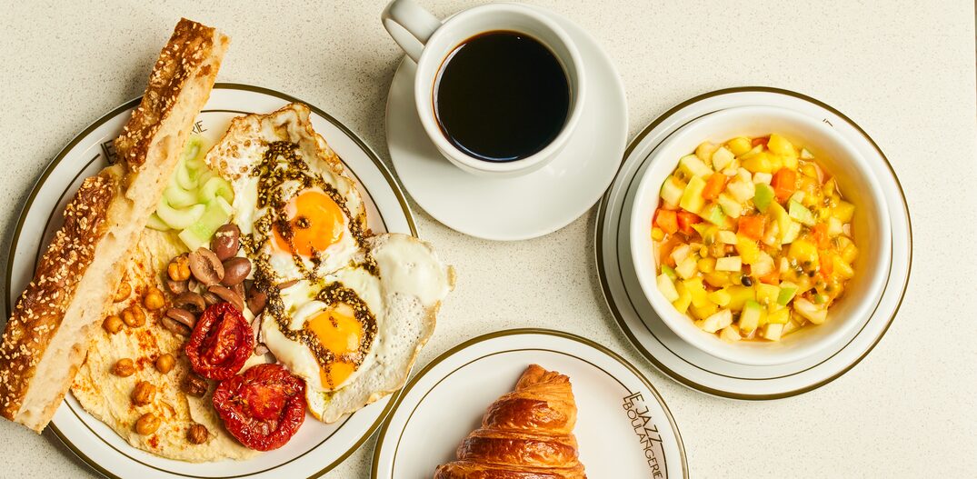 Pratos brancos com porções de pães e ovos fritos, salada de frutas, ovos com bacon, iogurte com granola e croissant + uma xícara de café preto. Foto: Laís Acsa