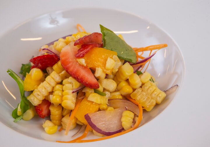 Salada de milho colorida, com morangos, cenoura, cebola roxa, rúcula,  servida em um prato fundo de cor branca, com bordas largas.