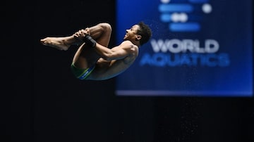 Isac Souza, um homem negro de cabelos curtos e vestindo apenas uma sunga, no ar segurando as canelas e projetando a cabeça em direção aos joelhos durante salto ornamental. A foto mostra o atleta de perfil. Foto: AFP