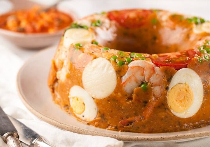 Cuscuz paulista, redondo, com massa alaranjada, ovos cozidos e camarão está sobre um prato de cerâmica de cor clara.