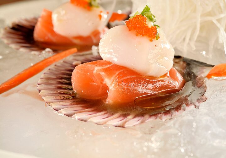 Duas conchas com pedaço de salmão, vieiras, ovas e folhinhas no topo. Ao lado das conchas, há gelo espalhado pela mesa.