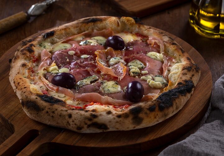 Pizza artesanal com presunto cru, azeitonas pretas e gorgonzola em cima de um suporte de madeira para pizza.