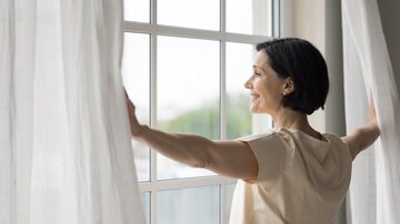 Side view rested middle-aged woman standing near window, open curtains, admires city view, enjoy peaceful, quiet morning, woke up early on weekend, welcoming, start new day, full of hope of happy day. Foto: fizkes/Adobe Stock          