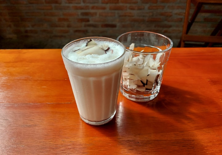 Sobre uma mesa de madeira, está um copo americano transparente contendo a batida de coco, enfeitado com lascas de coco. Atrás do copo, um copo transparente contendo lascas de coco.