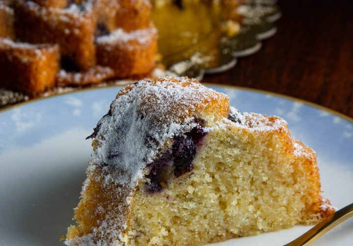 Fatia de bolo amarelo com pedaço de blueberry perto do topo e cobertura de açúcar. A fatia está sobre prato branco com borda azul-claro, ao lado de um garfo dourado. Ao fundo, mais fatias empilhadas e o bolo completo em prato dourado.