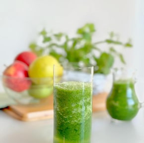 Em uma mesa branca está um copo alto e transparente com suco verde. Ao fundo, frutas. Foto: Edvânia Soares/Divulgação