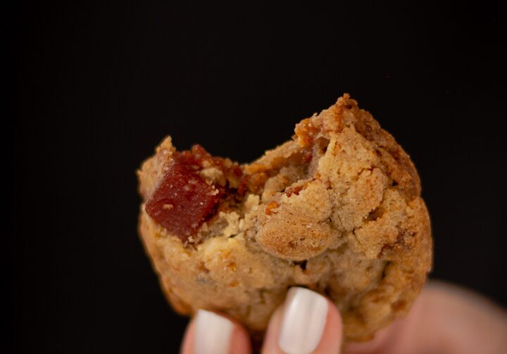Mão feminina com unhas brancas, segurando um cookie com goiabada, com uma mordida, revelando o recheio do meio. O fundo da foto é de cor preta.