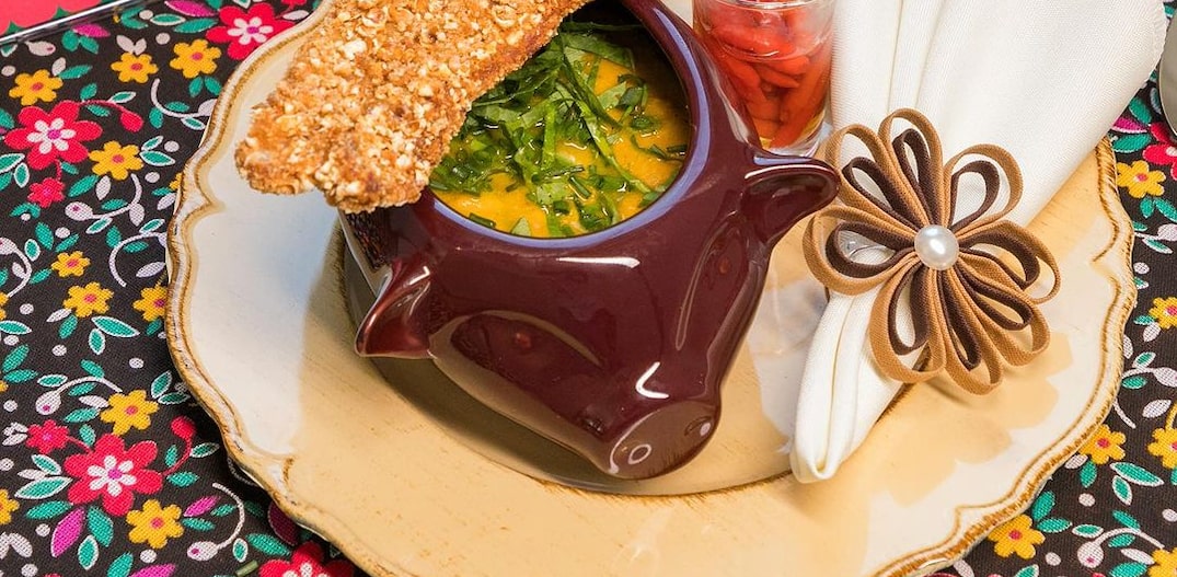 Arriê de abóbora servido em uma cumbuca, em cima de um prato raso de cor bege. O prato está em uma toalha de mesa florida, com uma placa rosa de letras brancas, com o nome da receita. Foto: Reprodução | Instagram @belohorizonte.mg