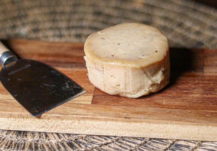 Sobre uma tábua de madeira, um queijo redondo e amarronzado nas beiradas está exposto, ele possui detalhes em preto, como temperos. Ao lado, uma espátula de corte de metal.