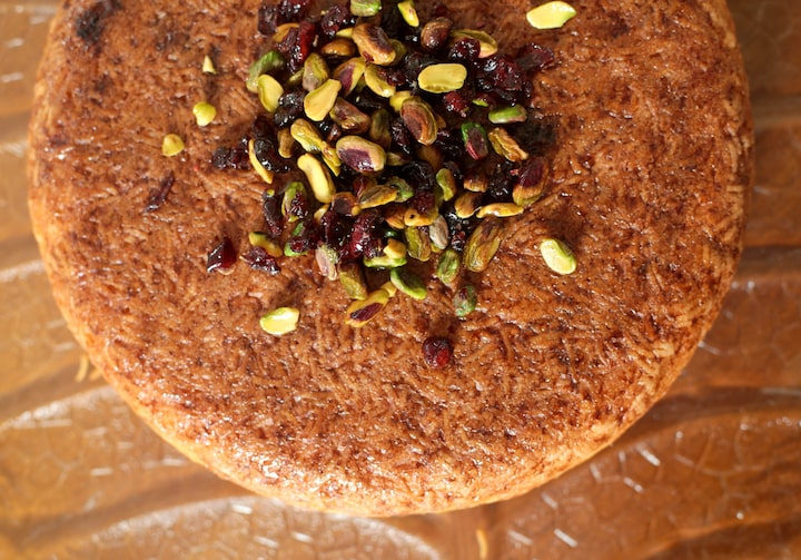 Sobre uma superfície marrom com relevos está o tah-chin, prato à base de arroz e frango, em forma de círculo - com aparência de um bolo em tom caramelo. Por cima, há pedaços de pistache e cranberry