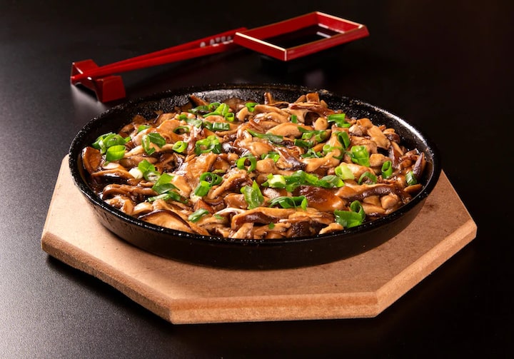 Um prato preto, em cima de uma superfície de madeira, está repleto de shitake com cebolinha. Atrás, há uma molheira preta e vermelha e dois hashi vermelhos num descanso. Os objetos estão sobre uma mesa preta