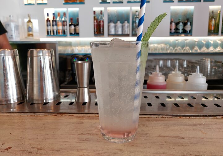 Sobre uma bancada de bar clara, está um copo transparente contendo um canudo branco com azul e o drinque Greek soda, uma soda italiana com xarope de rosas