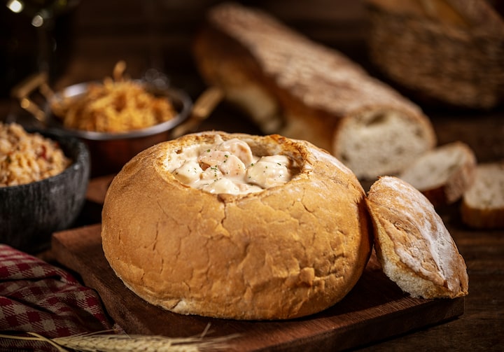 Em um pão italiano redondo, está o camarão aos quatro queijos enfeitado com salsinha por cima. Ao fundo, pão italiano, arroz provençal e batatas rústicas.