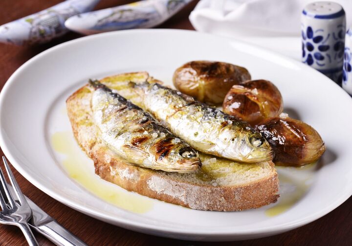 Em um prato raso branco, está um pedaço de broa com duas sardinhas grelhadas por cima e três batatas ao murro ao lado. Tudo regado com azeite.