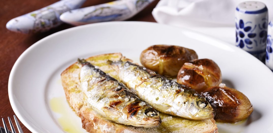 Em um prato raso branco, está um pedaço de broa com duas sardinhas grelhadas por cima e três batatas ao murro ao lado. Tudo regado com azeite. Foto: Rancho Português/Divulgação