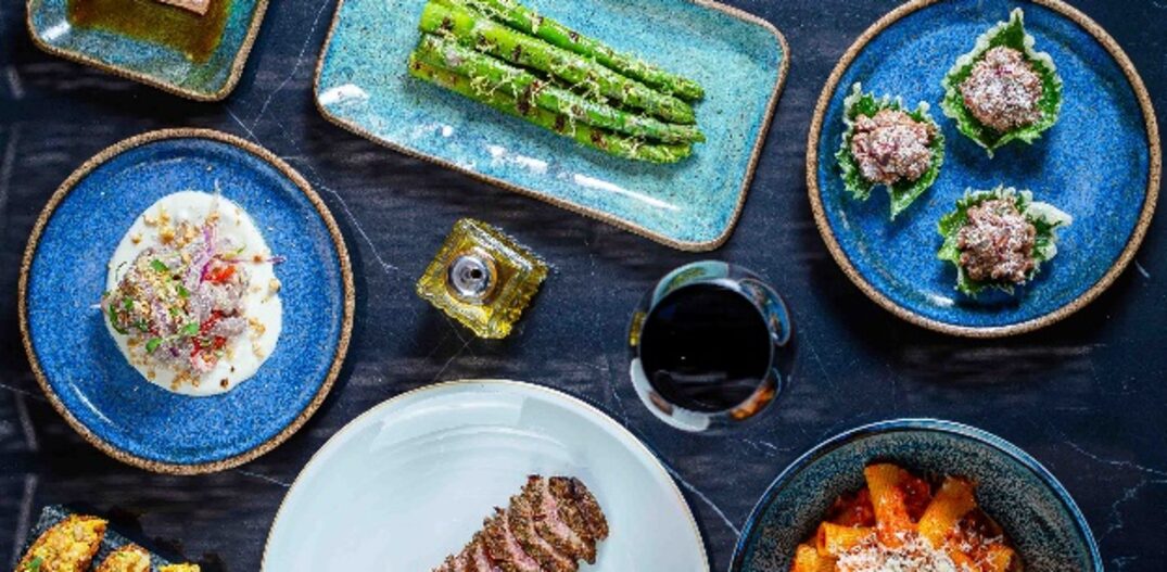 Variedade de pratos e utensílios de cozinha dispostos sobre uma mesa de cor azul escura. Os pratos servem variedades de carne fatiadas, macarrão rigatoni ao molho vermelho e queijo parmesão,. Foto: Greta von Hertwig