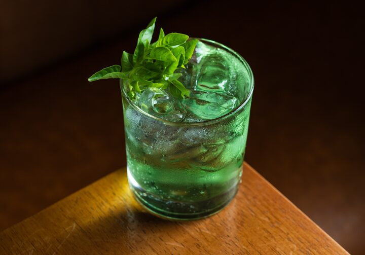 No canto de uma mesa de madeira, está um copo baixo com bastante gelo e contendo o drink em uma cor verde bem vibrante. O enfeite do drink fica a cargo de um ramo de manjericão.