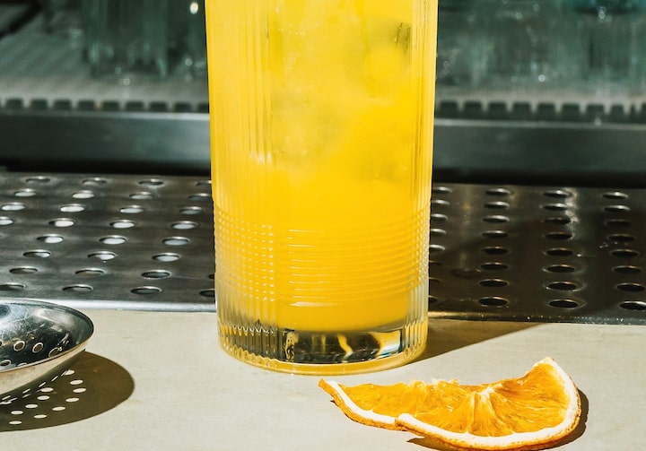 Em uma mesa de bar, laranja desidratada e um copo longo sobre a mesa. No copo, bastante gelo e o drinque bem amarelo.