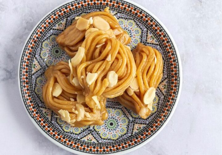 Prato estampado com massa em formato de espaguete e cobertura com pedaços de amêndoas. O prato está sobre mesa de mármore.
