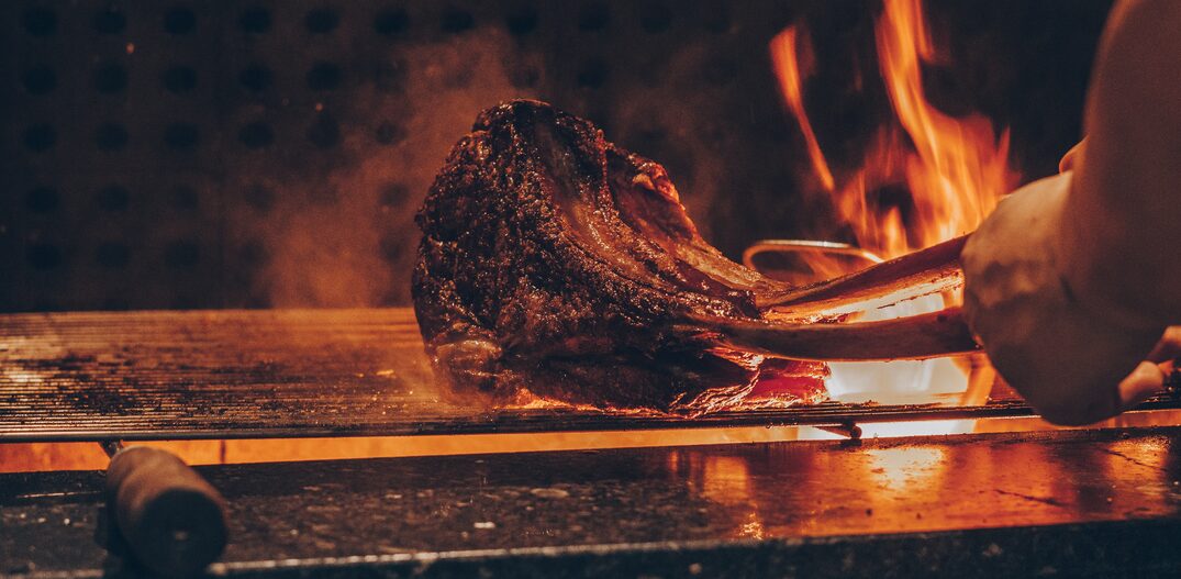 Mão de uma pessoa virando um pedaço de carne que estava na grelhada da churrasqueira. É possível ver o fogo subindo. Foto: Unsplash