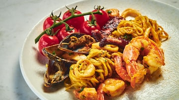 Em um prato raso branco, estão tomates pequenos, mexilhões, tentáculos de polvo, macarrão tipo espaguete e camarões. Foto: Rodolfo Regini/Divulgação