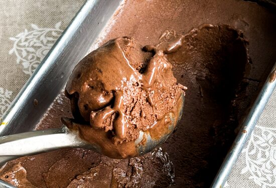 Colher de sorvete prateada pegando uma bola de sorvete de chocolate cremoso dentro de um pote de alumínio. O pote está sobre uma toalha branca rendada. Foto: Luciana Bonometti
