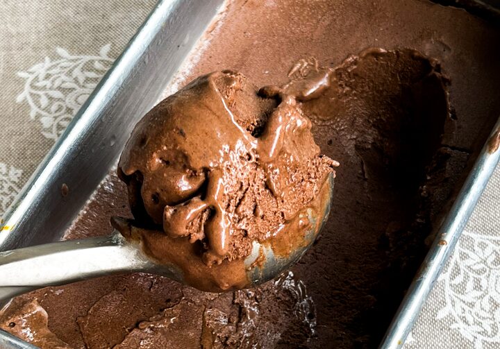 Colher de sorvete prateada pegando uma bola de sorvete de chocolate cremoso dentro de um pote de alumínio. O pote está sobre uma toalha branca rendada