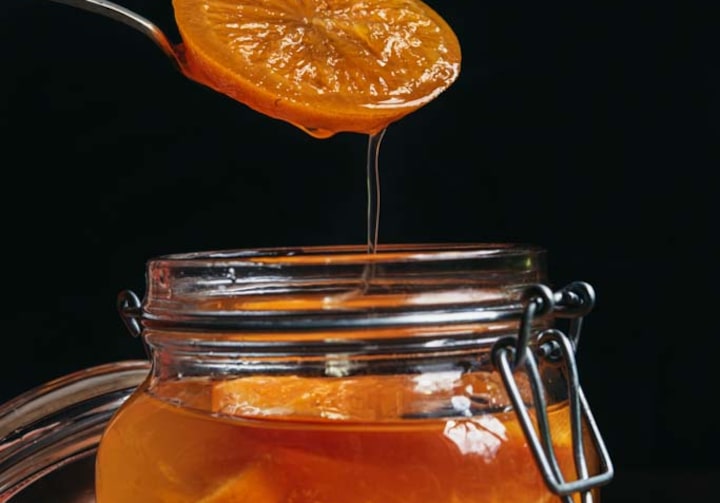 Compota de laranja da chef Helô Bacellar, servida em pote de vidro, com uma rodela de laranja da compota sendo suspensa por colher e despejando caldo dentro do pote.