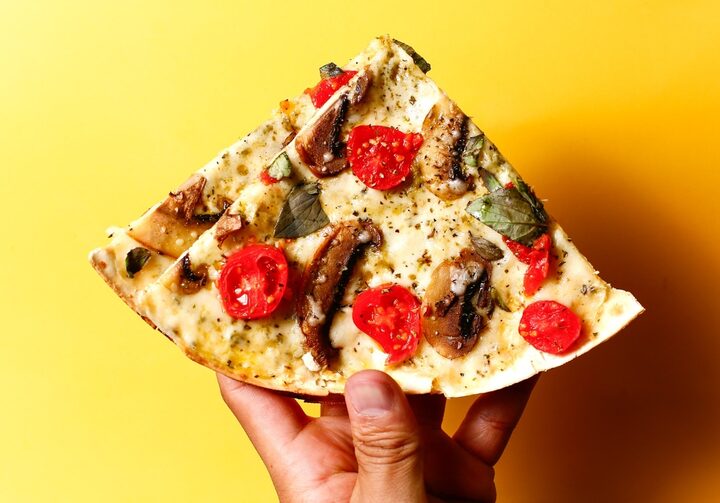 Uma fatia triangular de pizza com mussarela, cogumelos e tomates cereja fatiados, folhas de manjericão e orégano salpicado por cima é segurada por uma mão na frente de um fundo amarelo.