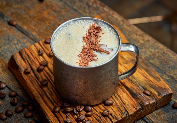 Em uma caneca de metal, está o drink Irish Coffee gelado, com raspas de chocolate enfeitando por cima e grãos de café dispostos na mesa de madeira onde está exposto.