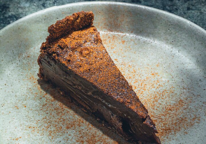 Sobre uma mesa de mármore cinza, um prato de bordas altas abriga uma fatia de torta de ganache polvilhada com chocolate em pó.