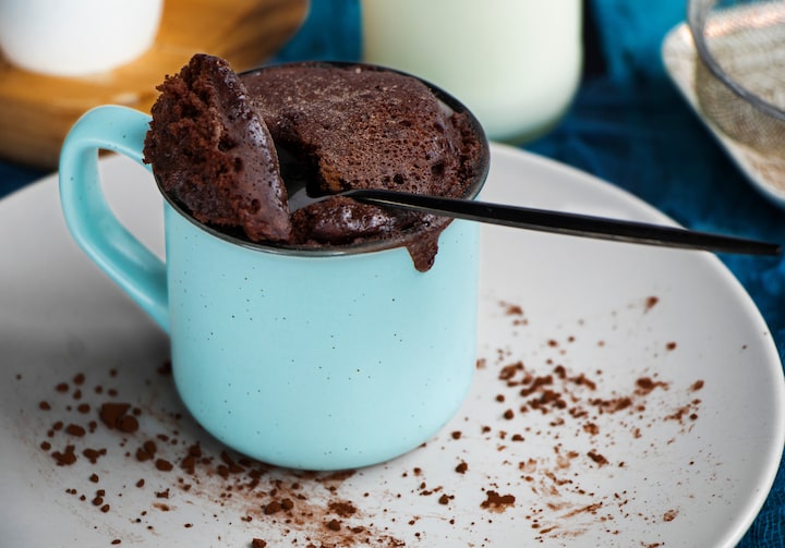 Brownie em uma caneca azul-clara e sobre um prato branco com chocolate em pó polvilhado. Uma colher retira um pedaço do bolo.