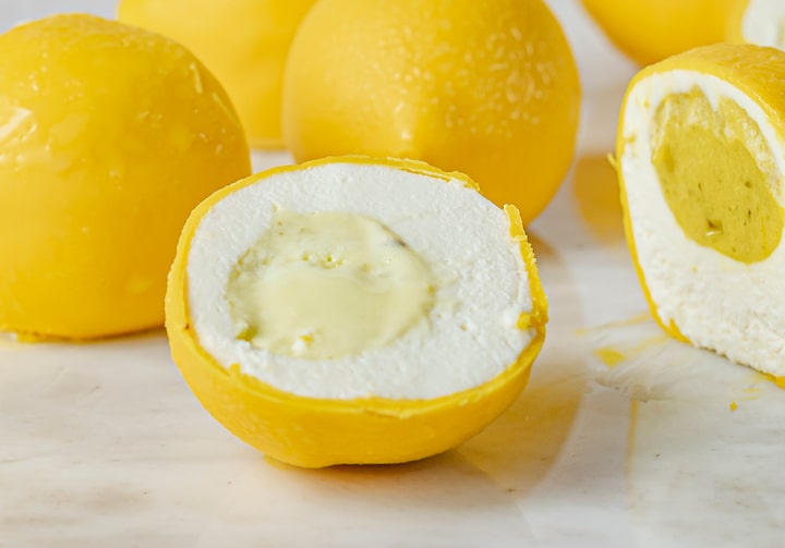 Sobre uma bancada branca, estão vários doces em formato de limão siciliano. Ao centro, um cortado pela metade expondo exterior amarelo forte e interior macio e branco, com um recheio cremoso e amarelado no meio.