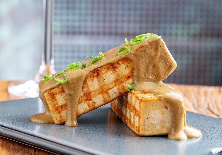 Sobre uma mesa de madeira, uma tábua de metal tem dois pedaços retangulares de tofu sobre ela. Grelhados com detalhes dourados quadriculados, os pedaços de tofu são enfeitados por um molho amarronzado claro e ramos verdes.
