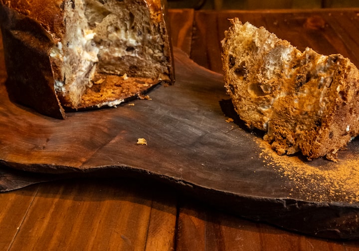 Em cima de uma tábua de madeira, está um chocotone cortado e recheado com um creme branco. Ao lado, há uma fatia do doce e cacau em pó polvilhado.