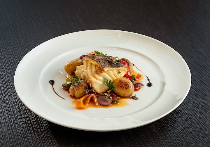 Sobre uma mesa de madeira escura, um bacalhau à napolitana está em cima de um prato fundo branco.