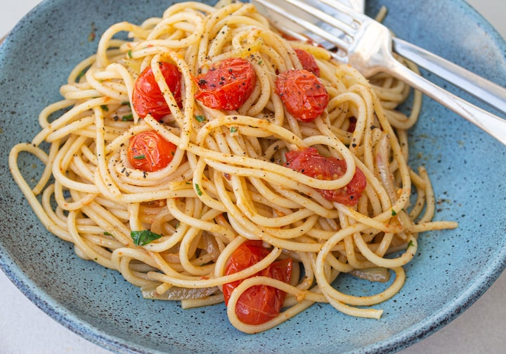 Espaguete, com tomates fatiados em cima, dentro de azul claro. O garfo e a colher estão tombados também dentro do prato