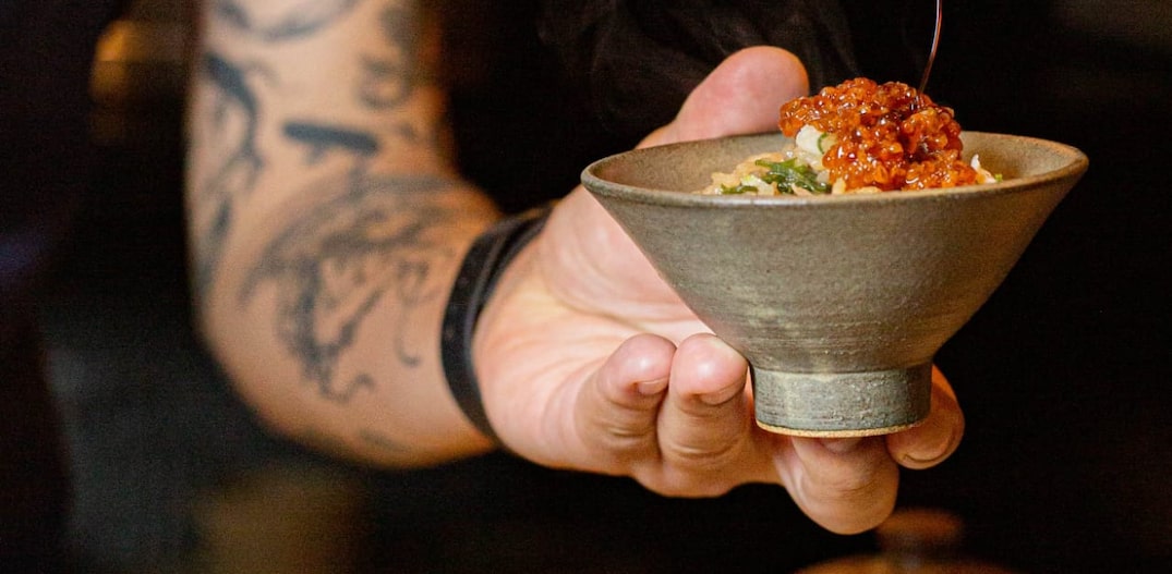 Braços e mãos de um homem com tatuagens. Nas mãos dele, um pincel sobre uma cumbuca de louça esverdeada com uma porção de comida japonesa. Foto: Tati Frison