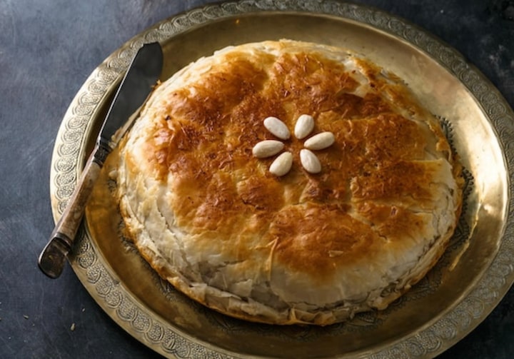 Prato dourado com torta marroquina, a bastela, decorada por amêndoas. Há uma faca prateada com cabo de marfim apoiada no prato, que está sobre uma superfície cinza chumbo.