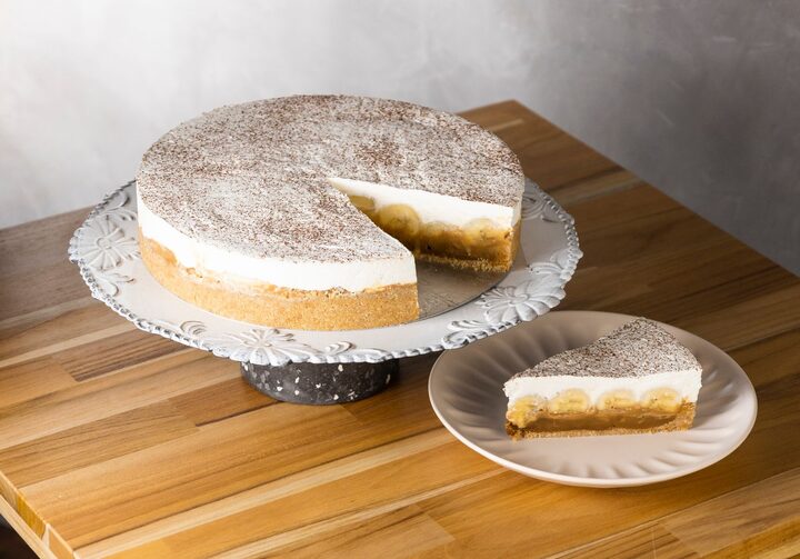 Banoffee, torta branca e bege sem uma fatia, em cima de um prato de bolo. Ao lado, uma fatia cortada em um prato pequeno. Tudo está sobre uma mesa de madeira. Fundo de parede cinza.