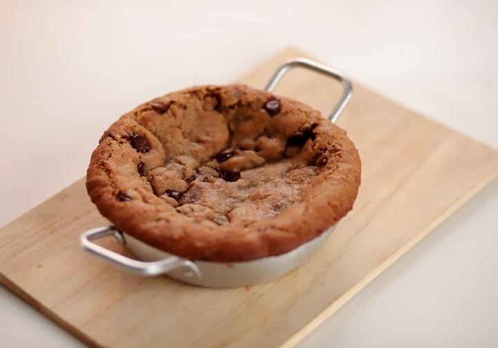 Em uma forma de alumínio com alças, está servida uma torta de cookie e gotas de chocolate, sobre uma tábua de madeira clara. A tábua está em cima de uma superfície bege.